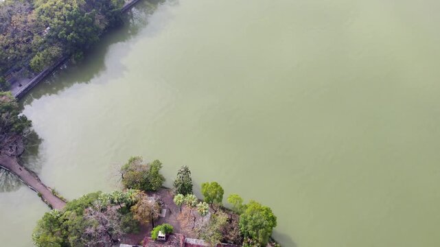 An aerial view of a tiny island with simple structures, connected by a narrow path, surrounded by calm greenish water and dense vegetation.