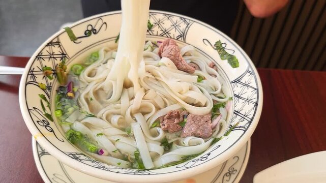 Chopsticks stir noodles and beef inside a bowl of pho soup, herbs and broth blending together.