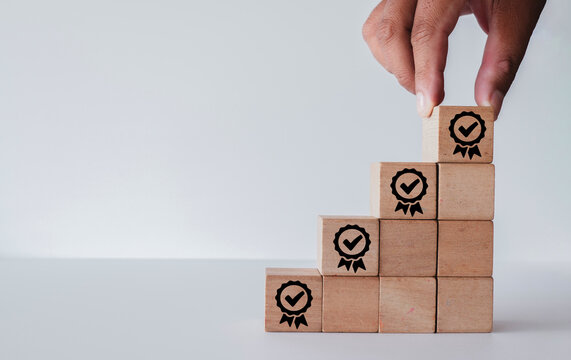 Close up of hand placing wooden block with quality guarantee badge icon on top of a staircase. Business concept for quality assurance, ISO certification, and standard improvement with copy space.