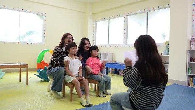 A young girl confidently reads aloud from a paper to a smiling, supportive audience of teachers and fellow students sitting together in a bright, colorful kindergarten classroom.