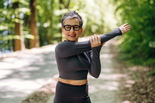Mature woman in black athletic wear stretches her arms while standing on a tree-lined path in a park during daylight, showcasing a healthy lifestyle and fitness routine