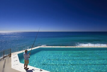 Naklejka premium Man fishing in ocean pool with clear blue sky and turquoise water.