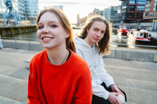Smiling friends sitting on step at urban harbor during sunset