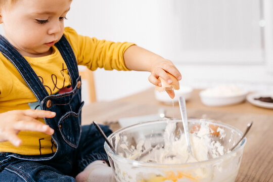 Toddler girl sitting on kitchen table throwing egg into bowl for preparing dough, partial view