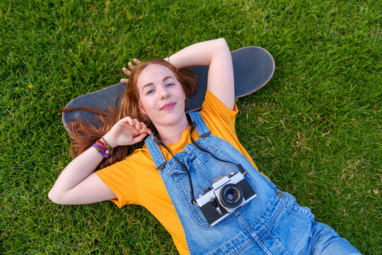 Smiling woman lying on grass with a skateboard and film camera