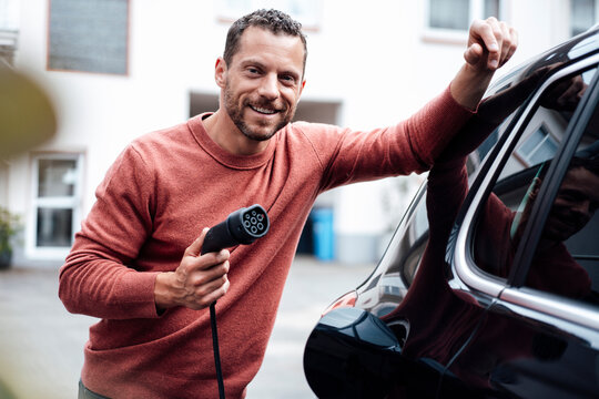 Smiling man holding charging cable leaning on electric car