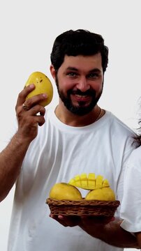 Indian young couple eating mango while standing isolated against white background, holding, promoting fresh seasonal aam or Alphonso happily, enjoying king of fruits with joyful expressions smiles
