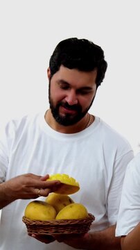 Indian young couple eating mango while standing isolated against white background, holding, promoting fresh seasonal aam or Alphonso happily, enjoying king of fruits with joyful expressions smiles