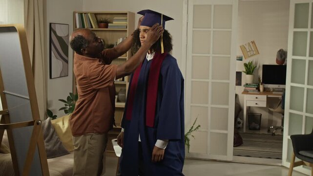 Medium long shot of African American middle aged father adjusting graduation cap on young adult son in living room before hugging him. For higher education, family support, student achievement promos