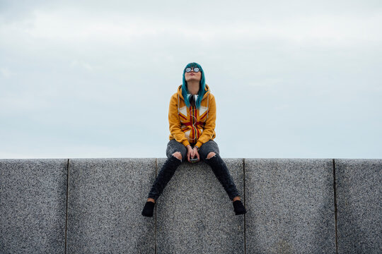 Young woman with dyed blue hair sitting on a wall looking up