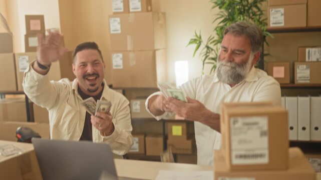 Man counting cash with hands and handing parcel near laptop amid stacked boxes in a small building; business celebration.