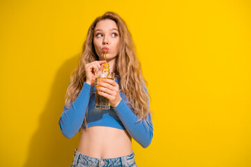 Smiling blonde girl in a blue top and jeans holding a glass on a vibrant yellow background showcasing stylish vibes.