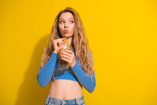 Smiling blonde girl in a blue top and jeans holding a glass on a vibrant yellow background showcasing stylish vibes.