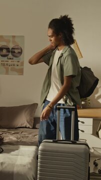 Vertical video of African American teenager picking up smartphone, putting on bags, and walking out of bedroom with suitcase. Clip is ideal for student travel or moving day commercials