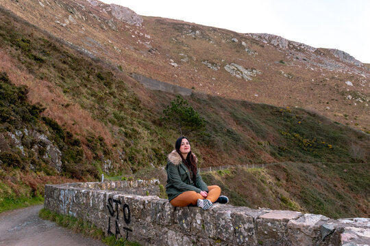 Young woman sitting cross-legged on a stone wall along a mountain trail in Ireland