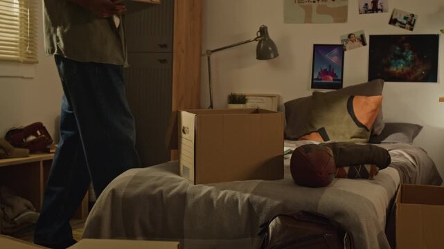 Handheld tilt up shot of Black teenager packing books into cardboard box in college dorm room. Clip is suitable for commercial videos about student relocation, campus life, and youth housing