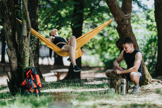 A hiking couple rests with one person lounging in a yellow hammock and the other unpacking a backpack. They relax under trees at a shady forest spot during an outdoor break.