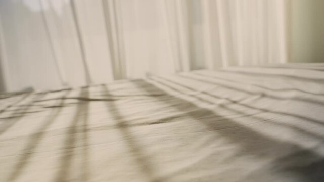 Soft sunlight casts striped shadows across a bed. White curtains gently filter the morning light. Blanket texture shows subtle folds and natural creases