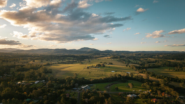 Aerial view of the rural landscape of Beechwood with rolling green pastures, scattered trees, and distant mountains under a soft golden hour sky in Beechwood, New South Wales, Australia.