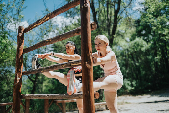 Two women perform leg stretches using a rustic wooden fence during an outdoor workout. A pair of athletes stretch their hamstrings and calves during a sunny park training session.