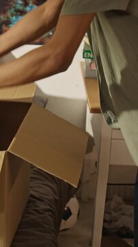Vertical video of African American teenage boy unpacking argyle sweater from cardboard box in bedroom. Footage ideal for student lifestyle, college moving, or second-hand fashion resale ads
