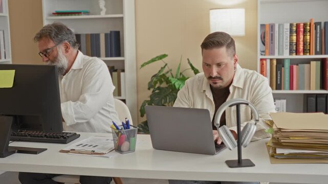 Two men at a desk, older man typing on a keyboard near monitor while younger man sorts stacked file folders beside a laptop in building; focused teamwork.