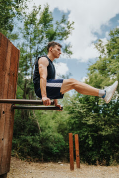 A fit young man performs an L-sit on parallel bars outdoors. He trains calisthenics on a wooden park apparatus under a blue sky.
