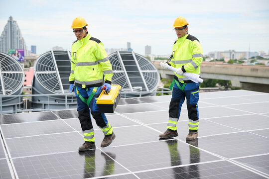 Technicians inspecting a solar panel array on a rooftop, dressed in safety gear. One is holding a blueprint and the other a toolbox. They appear focused on their work