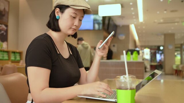 An adult asian woman uses a smartphone while operating a laptop at a table in an indoor public space. A drink is placed beside her.