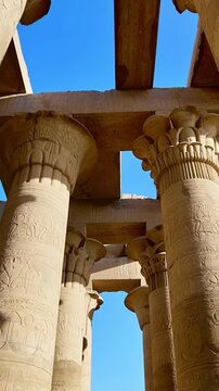 Kom Ombo temple columns with composite capitals and hieroglyphic carvings viewed from below against blue sky in Egypt, upward perspective of ancient sandstone architecture