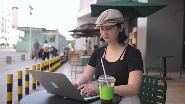 An asian adult woman sits at an outdoor cafe table, using a laptop while drinking a beverage with a straw.