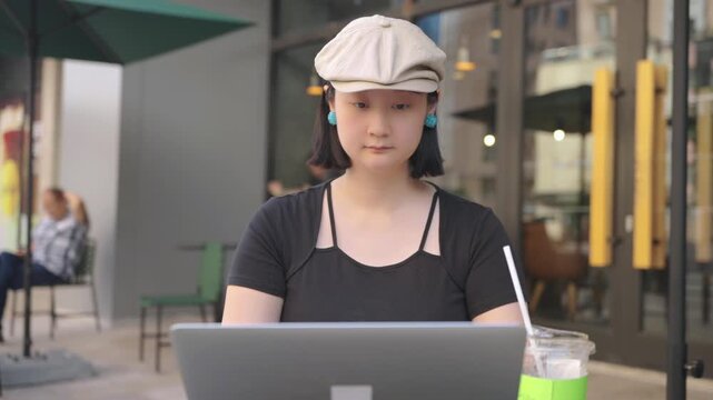 An adult Asian woman sits at an outdoor cafe table, using a laptop with a green drink beside her, in a daytime urban setting.