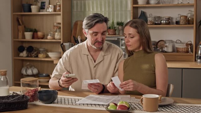 Medium sot of middle aged Caucasian couple sitting in kitchen, checking paper receipts and using smartphone for home budgeting. Footage of financial planning apps or banking services commercials