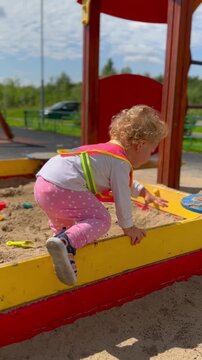 Child playing in sandbox at playground. Toddler wearing safety vest climbing over wooden barrier. Active childhood game outdoors in summer sunny park environment.