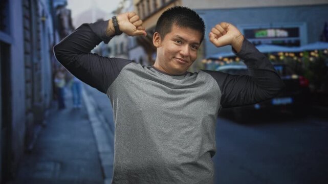 Man raises arms and adjusts shirt collar on a narrow street, smiling with eyes half closed while lifting hands above head in a stretch; playful confidence.