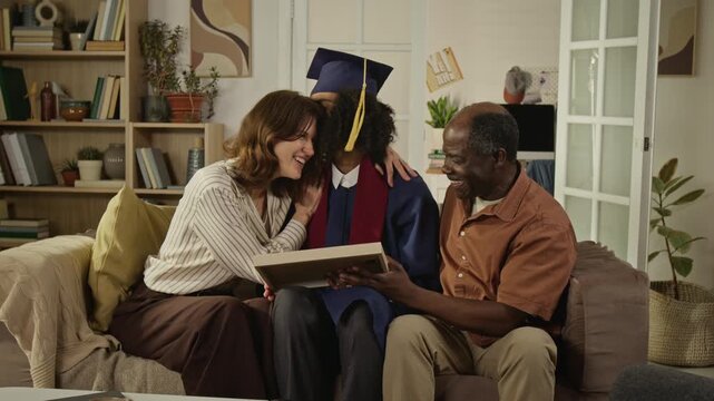 Medium long shot of happy diverse family with middle aged Caucasian mothe, Black father and teenage graduate sitting on sofa, celebrating achievement. Ideal for higher education marketing campaign