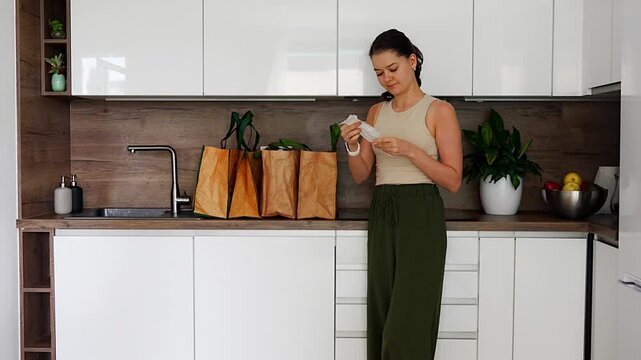 Woman checking grocery receipt after food delivery in reusable bags. Budget control and online shopping concept