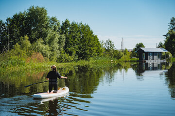 Fototapeta premium Person paddling standup paddleboard along calm river beside tree lined shore, reeds, reflections, and small docked cabin in peaceful summer landscape under bright daylight