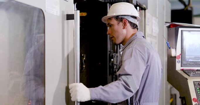 Asian man engineer slides CNC machine door while reviewing internal workspace, performing equipment check and safe access procedure for precision manufacturing operation in industrial plant facility