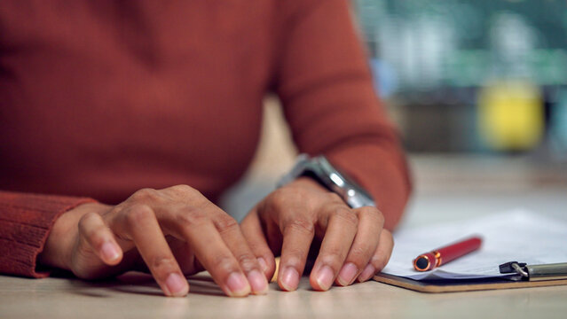 Hands rest on a desk with a pen nearby. Person wears a warm-toned sweater and a watch. Fingers are slightly tense, suggesting concentration or thought