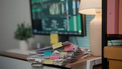 Naklejka premium Stacked papers with colorful sticky notes sit on a desk. Computer monitor displays financial charts in green and black. Soft lamp casts warm light beside the screen