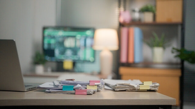 Focused work scene with stacked papers and sticky notes. Laptop rests beside a bright monitor displaying charts. Soft lamp casts warm glow across the workspace