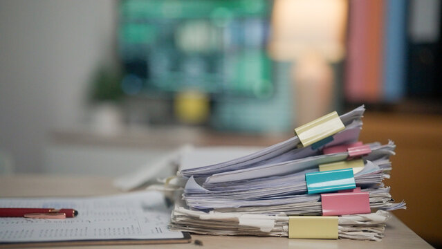 Stack of papers marked with colorful tabs rests on a desk. Red pen lies nearby, suggesting recent work or planning. Background shows a blurred computer screen and warm lamp light