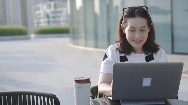 An adult woman uses a laptop while seated outdoors in an urban plaza, with a coffee cup nearby. She wears casual clothing and has sunglasses on her head, appearing focused and relaxed.
