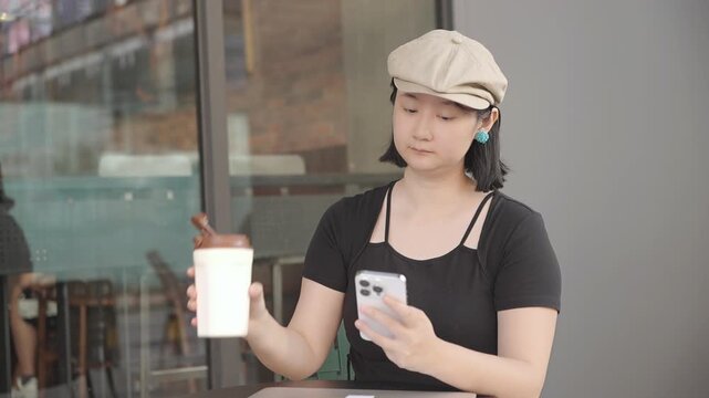 An young asian woman sits at an outdoor cafe table, holding a coffee cup and using a smartphone, wearing a beige beret and black top.