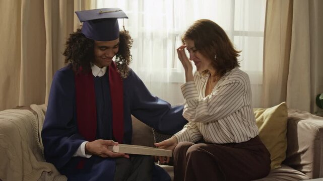 Medium long shot of Black teenager in graduation gown comforting crying Caucasian middle aged mother holding certificate on sofa. Footage ideal for university enrollment promos, family milestone ads