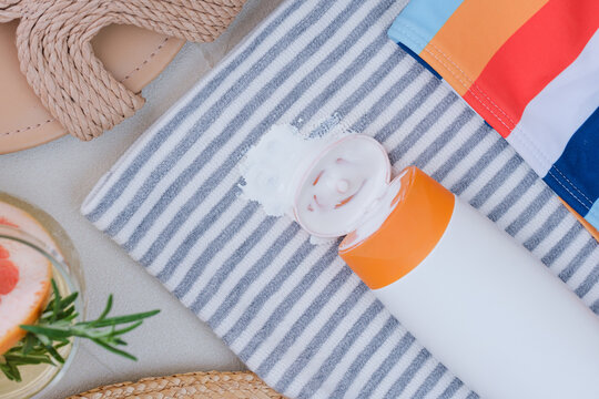 Sunscreen spilled on a towel with stripes. A drink with a slice of grapefruit and herbs sits nearby. Sandals are visible on the side, showing a relaxed beach scene. top view