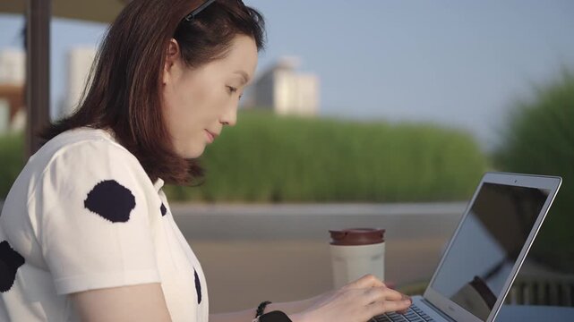 A woman works on a laptop at an outdoor urban patio table, with a drink and greenery around on a sunny day.