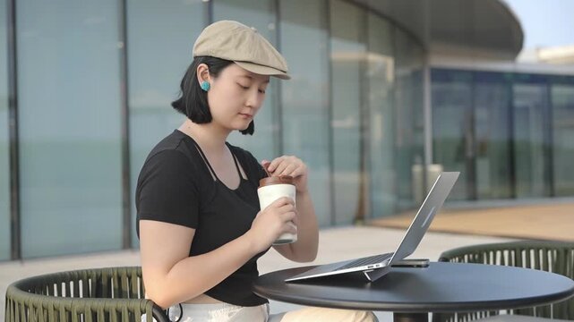 A young asian woman works on a laptop at an outdoor round table, with a coffee cup, in front of a modern glass building.
