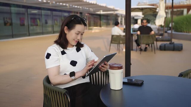 An adult asian woman uses a tablet while seated at an outdoor cafe table with a coffee cup nearby, in a bright daytime setting.